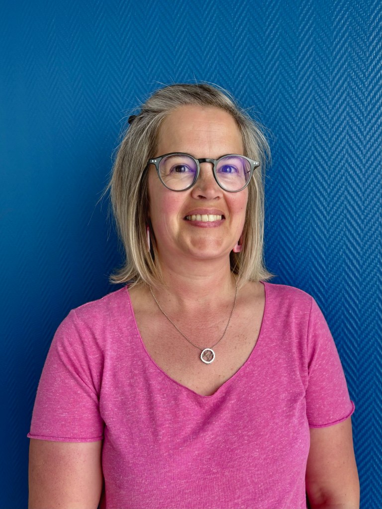 Portrait d'une femme souriante portant un t-shirt rose et des lunettes, devant un fond bleu texturé.
